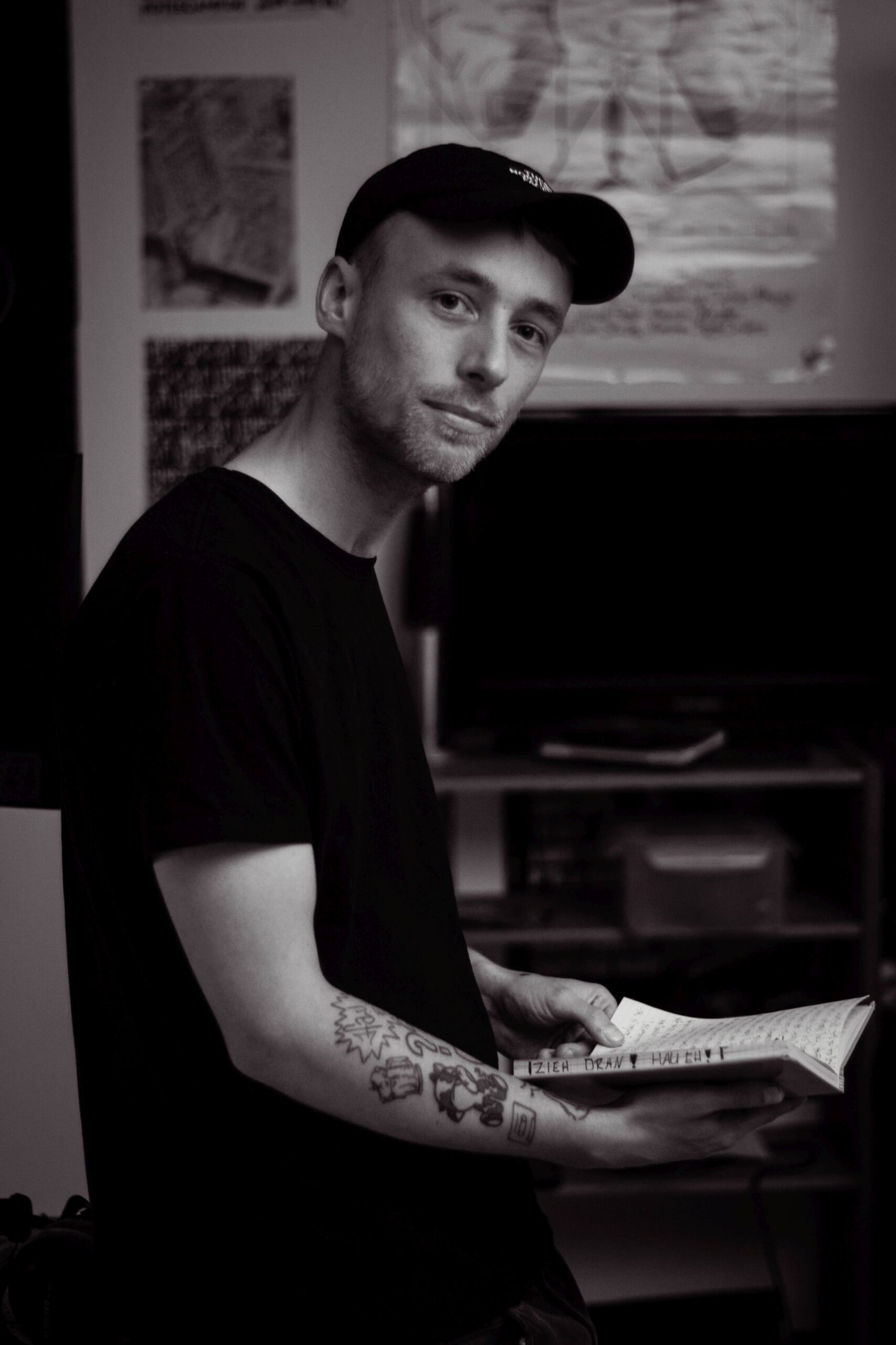 Rapper Adrinalin sitzt in seinem Studio auf der Kante des Tisches. Er sitzt seitlich nach rechts und sieht zur Kamera. In der Hand hält er ein geöffnetes Notizbuch mit seinen Rap-Texten. Adrinalin trägt eine schwarze Kappe und ein schwarzes T-Shirt.