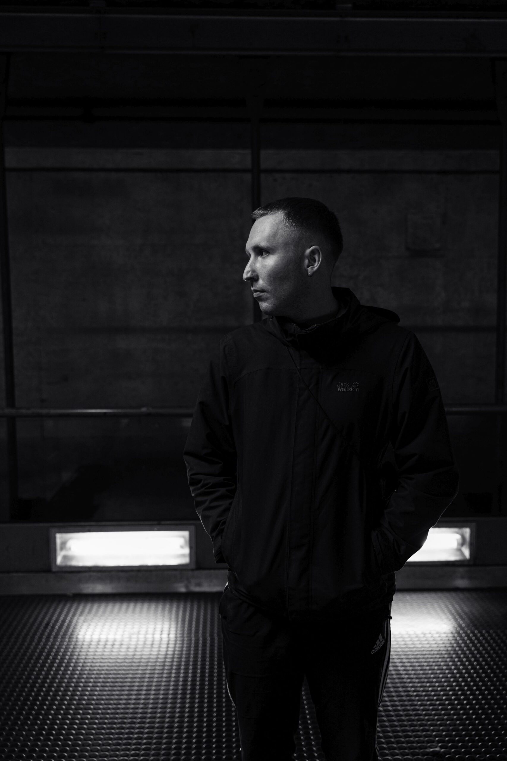 Rappe DrikOne steht in einem U-Bahnhof. Er steht seitlich zur Kamera, sieht nach links und trägt schwarze Kleidung ohne Kopfbedeckung.