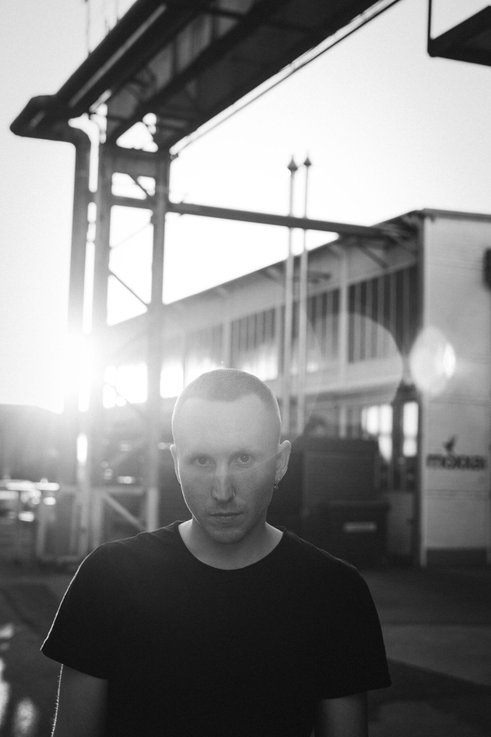 Rapper DrikOne steht im bei Sonnenuntergang im Gegenlicht und sieht in die Kamera. Er trägt keine Kappe und ein schwarzes T-Shirt.