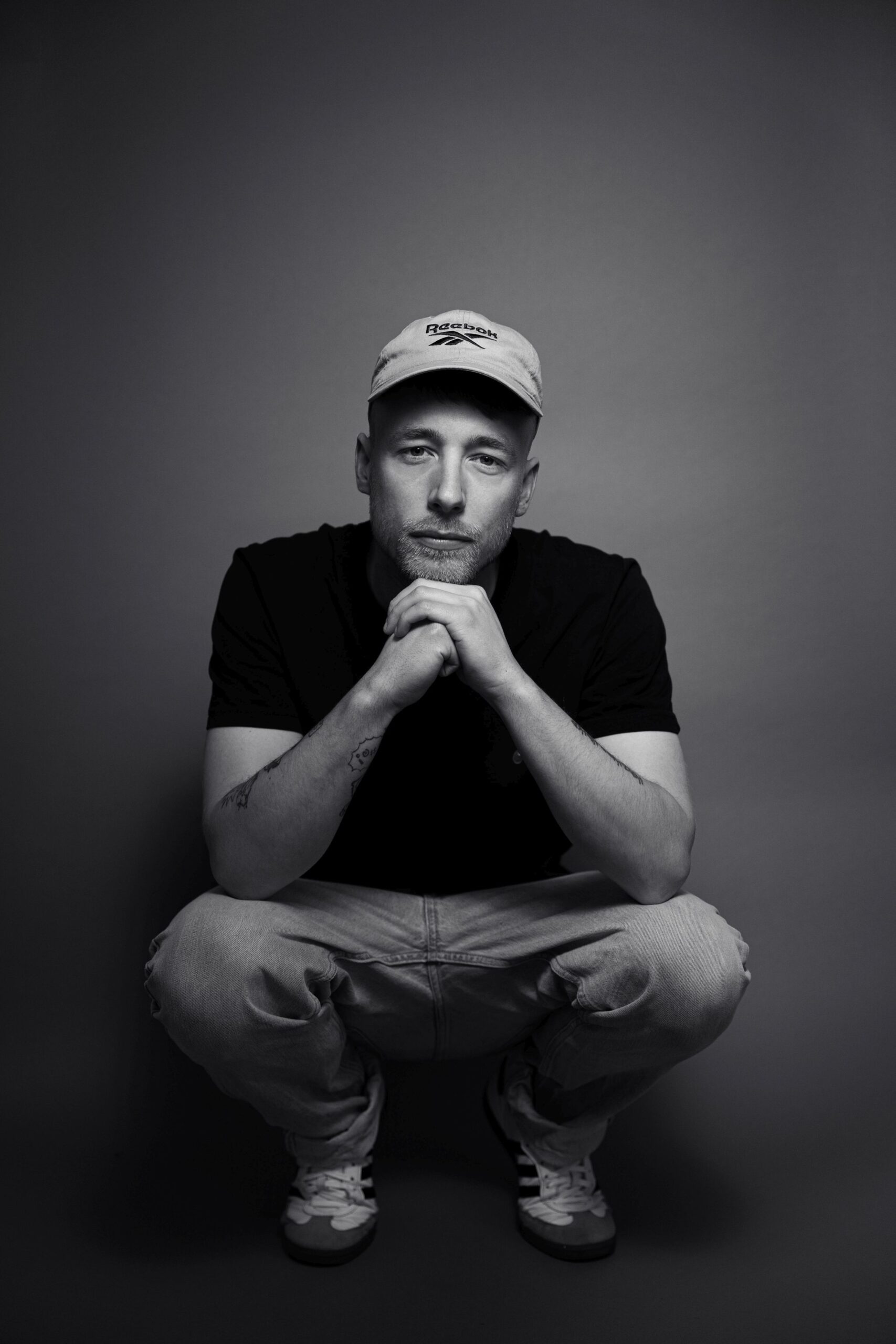 Rapper Adrinalin sitzt in der Hocke vor der Kamera. Er hat seine Ellenbogen auf die Oberschenkel gestützt und seine Hände stützen sein Kinn. Er trägt ein schwarzes T-Shirt und eine graue Kappe. Außerdem eine Jeans und weiße Turnschuhe. Er hock vor einem dunkelgrauen Hintergrund. Das Bild ist eine Schwarz-weiß-Aufnahme.