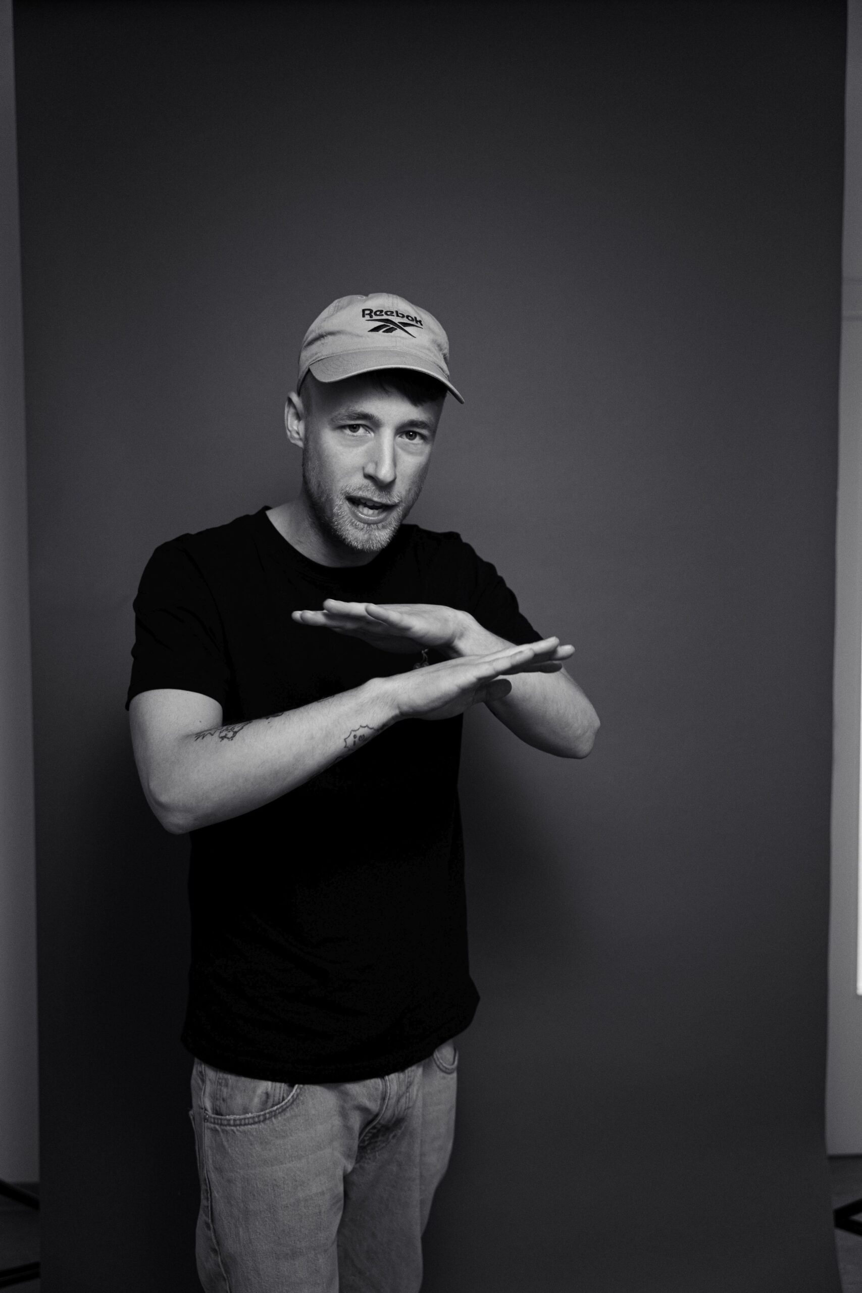 Rapper Adrinalin steht vor einem dunkelgrauen Hintergrund. Mit eine Geste kreuzt er seine Arme, denn er wurde beim Rappen fotografiert. Er sieht dabei in die Kamera. Er trägt eine graue Kappe, ein schwarzes T-Shirt und eine helle Jeans.