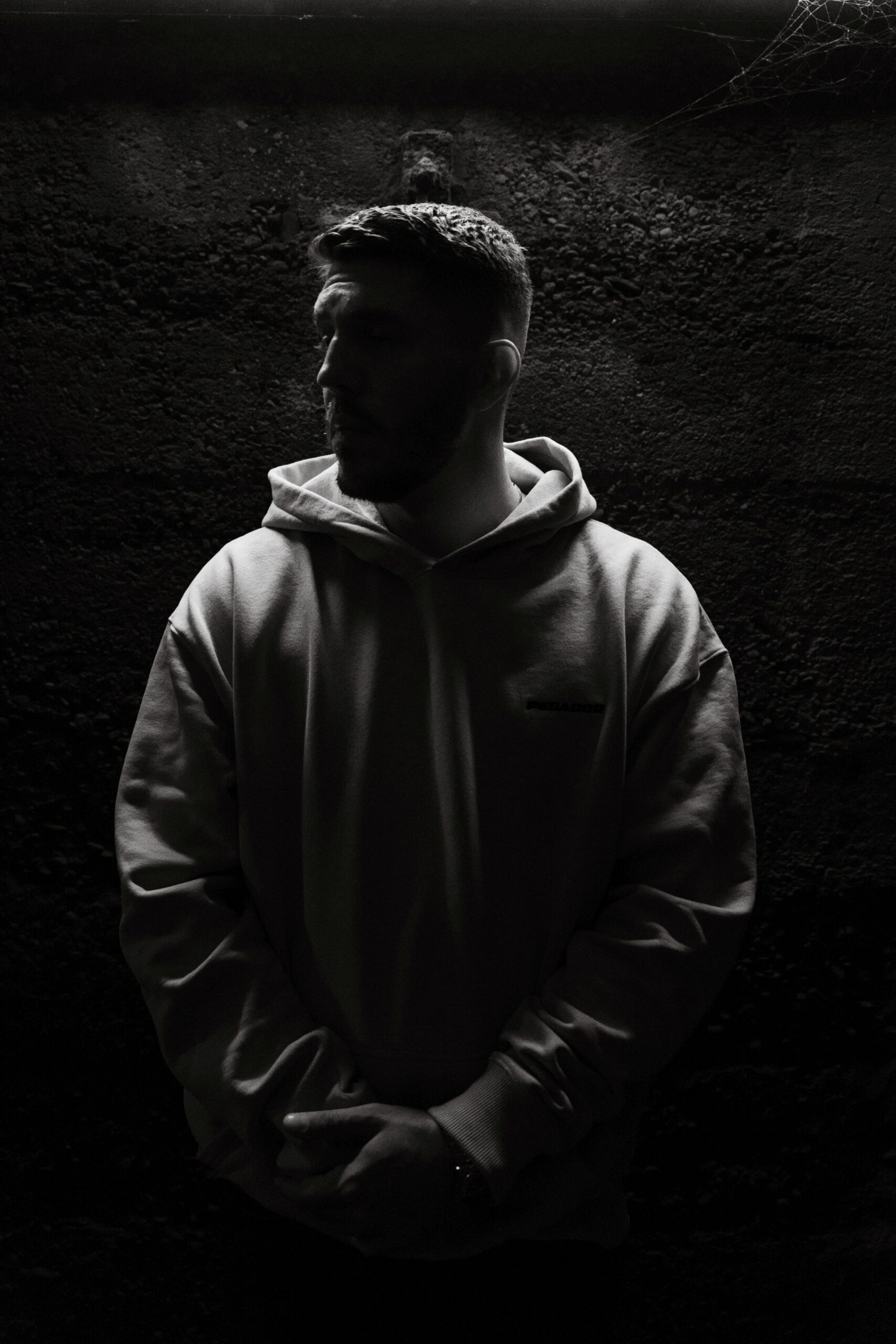 Der Rapper IInkasso aus Oberhausen steht vor einer Steinwand. Das Licht scheint über seinem Kopf und beleuchtet nur den Hinterkopf und die Schultern. Sein Gesicht liegt im Dunkeln. Inkasso sieht zur recht Seite und hält seine Hände. Das Foto wurde von Fotograf Toni Borgert aufgenommen.