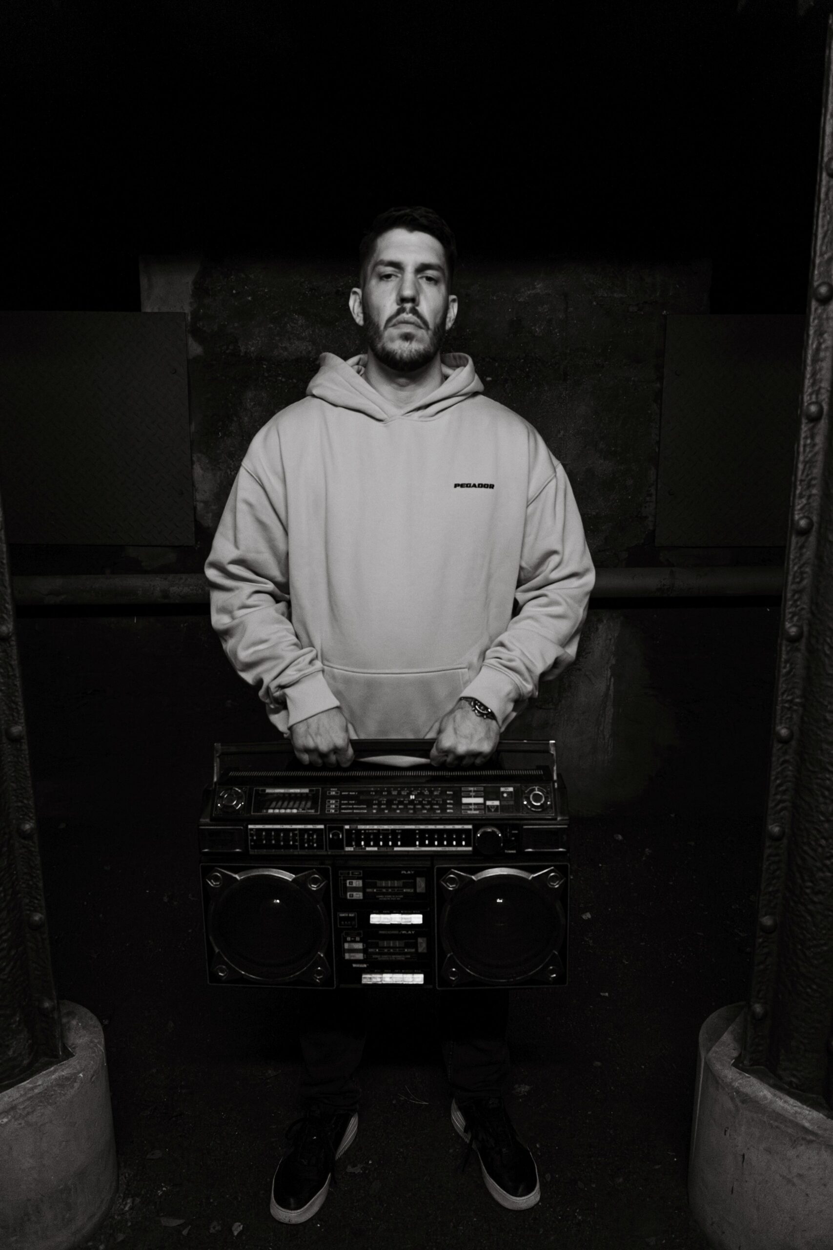 Der Rapper Inkasso aus Oberhausen steht vor einer Steinwand in einem Tunnel. Er trägt einen hellen Pullover und hält ein großes Radio in beiden Händen. Dabei sieht er mit einem ernsten Gesichtsausdruck in die Kamera. Das Foto wurde von Fotograf Toni Borgert aufgenommen.