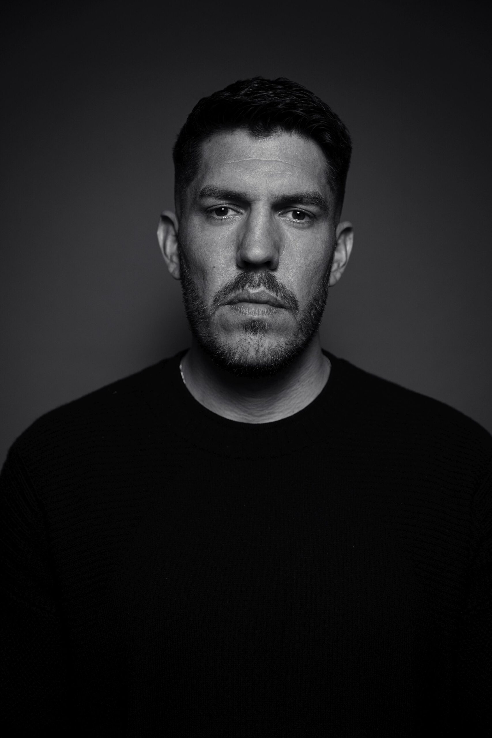 Der Rapper Inkasso aus Oberhausen steht vor einem grauen Papierhintergrund. Man sieht nur seinen Oberkörper von der Brust zum Kopf. Er trägt einen schwarzen Pullover und sieht mit ernstem Gesichtsausdruck in die Kamera. Das Foto wurde von Fotograf Toni Borgert aufgenommen.