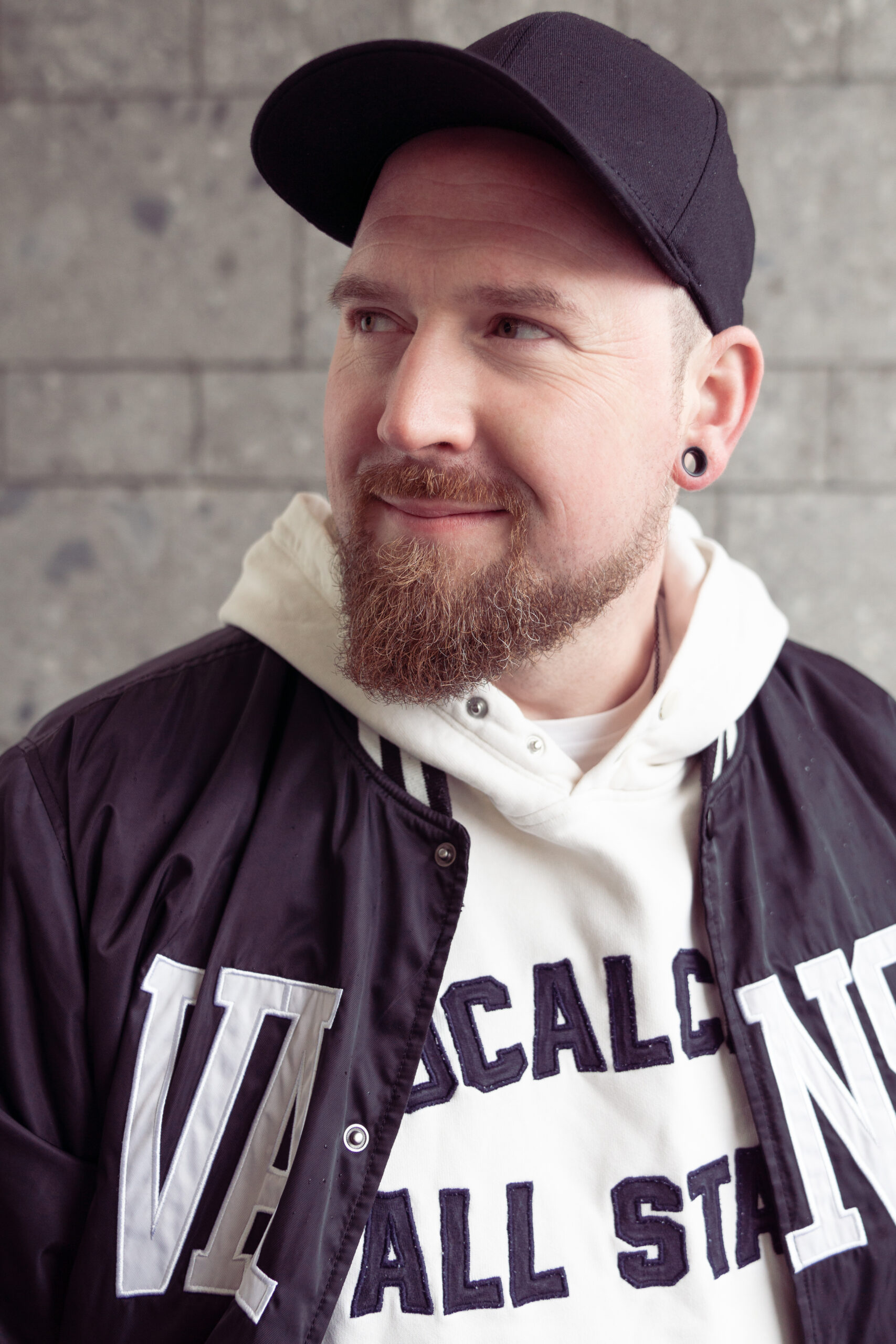 Das Foto zeigt einen Mann mit Baseball-Kappe und Baseball-Jacke. Darunter trägt einen Kapuzenpullover mit einer Aufschrift, die von der Jacke verdeckt wird. Er trägt außerdem einen Bart und lächelt sanft. Er sieht nach links an der Kamera vorbei. Das Foto wurde von Fotograf Toni Borgert aufgenommen.