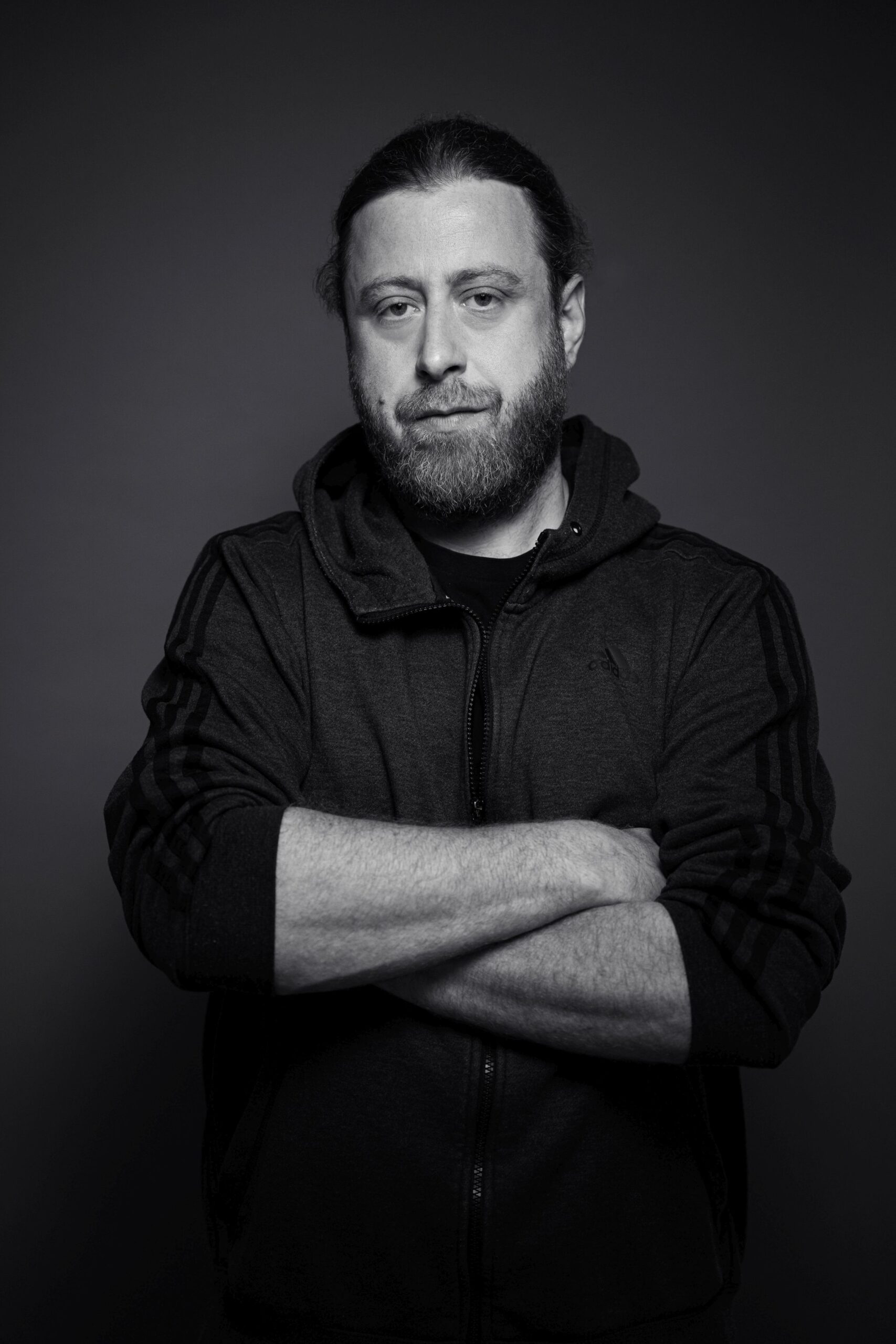 Das Bild zeigt den Rapper Keeko aus dem Ruhregebiet in NRW. Er steht vor einem grauen Papierhintergrund und trägt einen dunklen Pullover mit Kapute. Er hat die Arme vor sich verschränkt und sie mit geschlossenem Mund und ernstem Gesichtsausdruck in die Kamera. Das Foto wurde von Fotograf Toni Borgert aufgenommen.