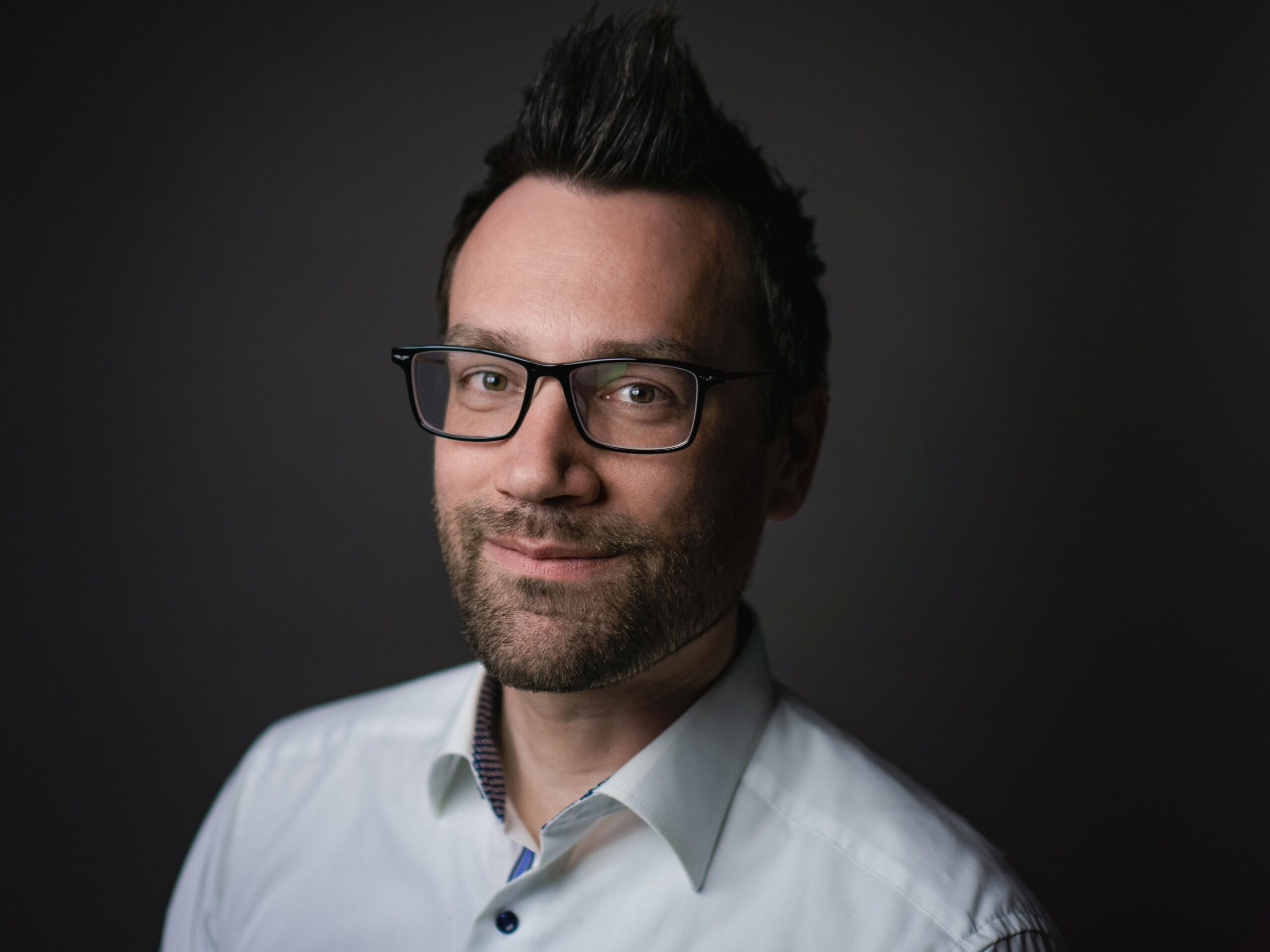 Das Foto zeigt eine männliche Person im Business-Look. Sie trägt ein Hemd mit Knöpfen. Der erste Knopf am Hals ist geöffnet. Die Person hat dunkle Haare, die leicht mach oben gegelt sind. Sie sitzt seitlich nach links gerichtet und sieht in die Kamera und lächelt. Das Foto wurde in einem Studio aufgenommen. Der Fotograf heißt Toni Borgert.