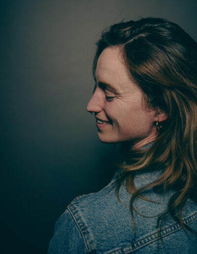 Das Bild zeigt eine Frau von der Seite die über ihre linke Schulter zur Seite sieht. Zu sehen ist ihre linke Gesichtshälfte und sie lächelt. Sie hat schulterlange blonde Haare und trägt eine Jeansjacke