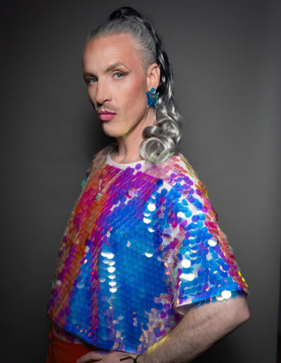 Das Bild zeigt eine nicht-binäre Person mit grauen Haaren die zum Zopf gebunden sind. Die Person steht seitlich zur Kamera und sieht mit einem überlegenen Blick und gehobener linker Augenbraue in die Kamera. Die Person trägt eine Paillettenbluse deren Farben orange, blaut und violet changieren. Das Bild wurde von Fotograf Toni Borgert aufgenommen.