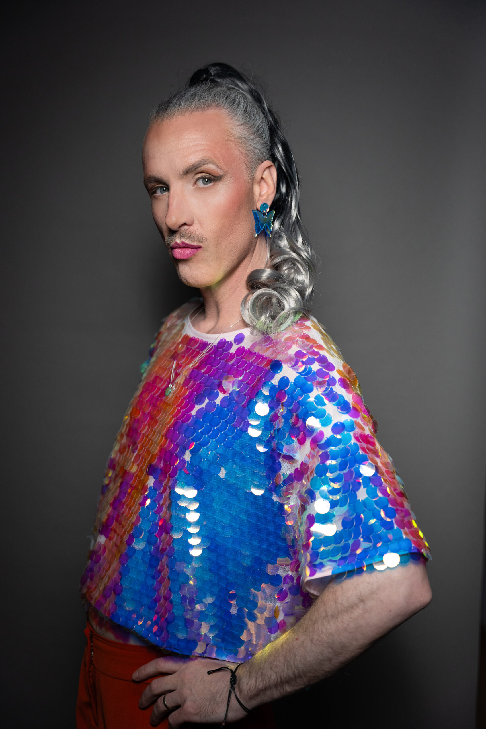 Das Bild zeigt eine nicht-binäre Person mit grauen Haaren die zum Zopf gebunden sind. Die Person steht seitlich zur Kamera und sieht mit einem überlegenen Blick und gehobener linker Augenbraue in die Kamera. Die Person trägt eine Paillettenbluse deren Farben orange, blaut und violet changieren. Das Bild wurde von Fotograf Toni Borgert aufgenommen.