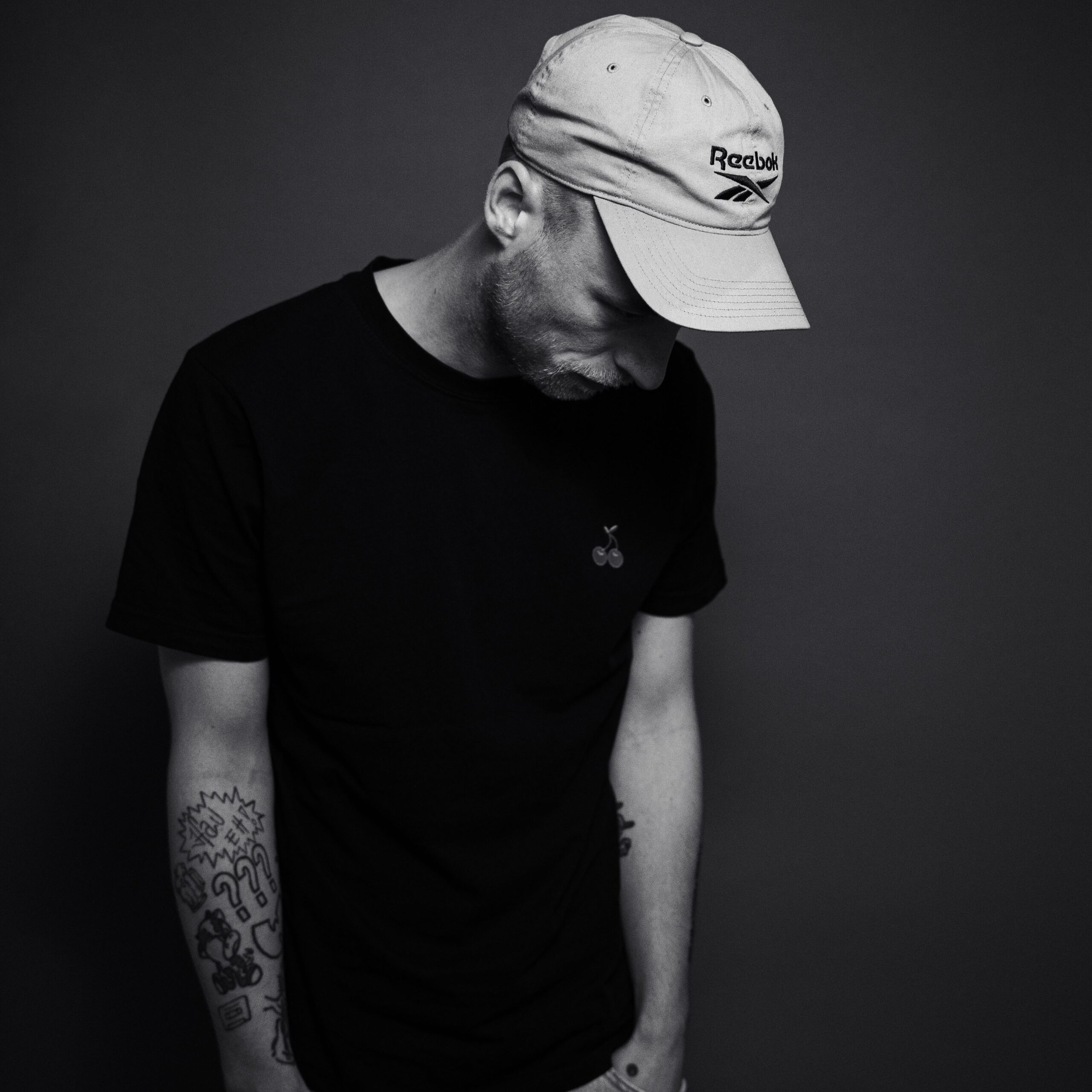 Das Schwarz-Weiß-Foto zeigt den Rapper Adrinalin. Er hat die Hände in den Taschen und sieht seitlich nach unten. Er trägt eine graue Kappe und ein schwarzes T-Shirt. Das Foto ist das Cover zur Single "Defizit". Es wurde von Fotograf Toni Borgert aufgenommen.