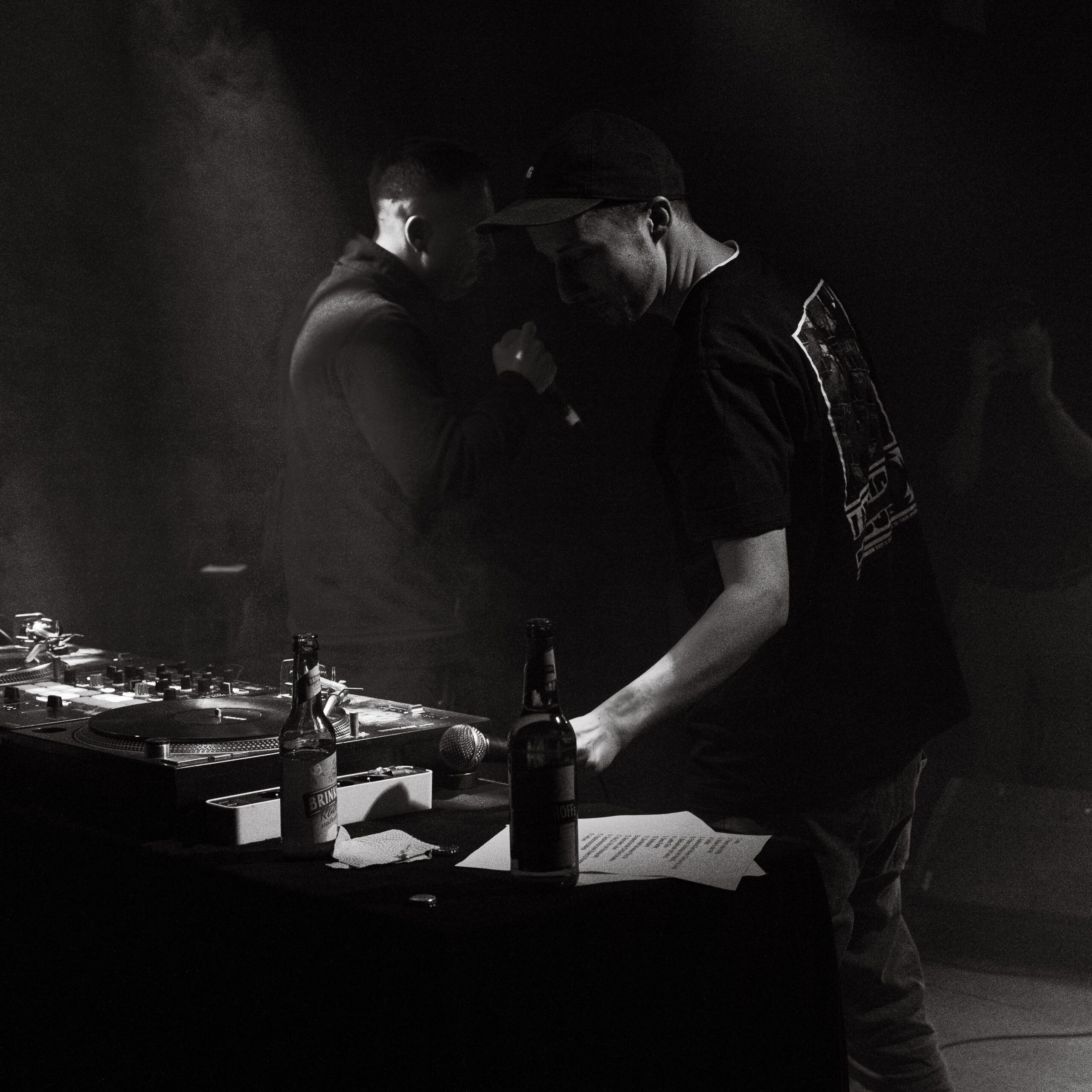 Das Schwarz-Weiss-Foto zeigt die Rapper DrikOne und Coba bei einem Konzert. Sie stehen voreinander und Coba verdeckt DrikOne. Die beiden stehen auf einer Bühne. Im Vordergrund steht ein DJ-Pult und es sind Bierfalaschen zu erkennen. Das Bild von Fotograf Toni Borgert aufgenommen.