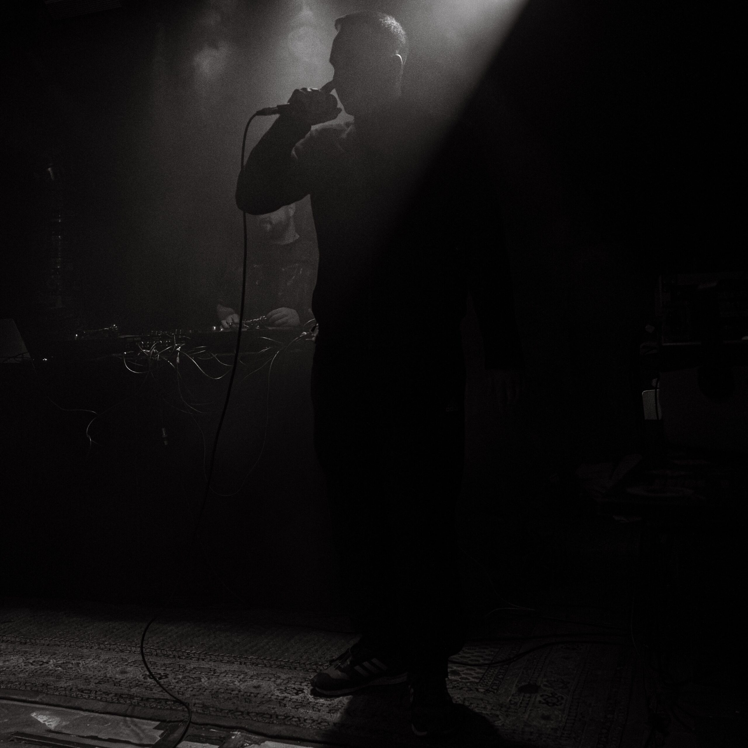 Das Foto zeigt den Rapper DrikOne. Er steht auf der Bühne und wird von hinten von einem Licht angestrahlt. Es ist nur der Umriss des Rappers zu erkennen. Drikone hat ein Mikrofon in der rechten Hand und trägt dunkle Kleidung. Das Licht ist diffus und eine Nebelmaschine verstärkt diesen Effekt. Es handelt sich um ein Schwarz-Weiss-Foto das von Fotograf Toni Borgert aufgenommen wurde.