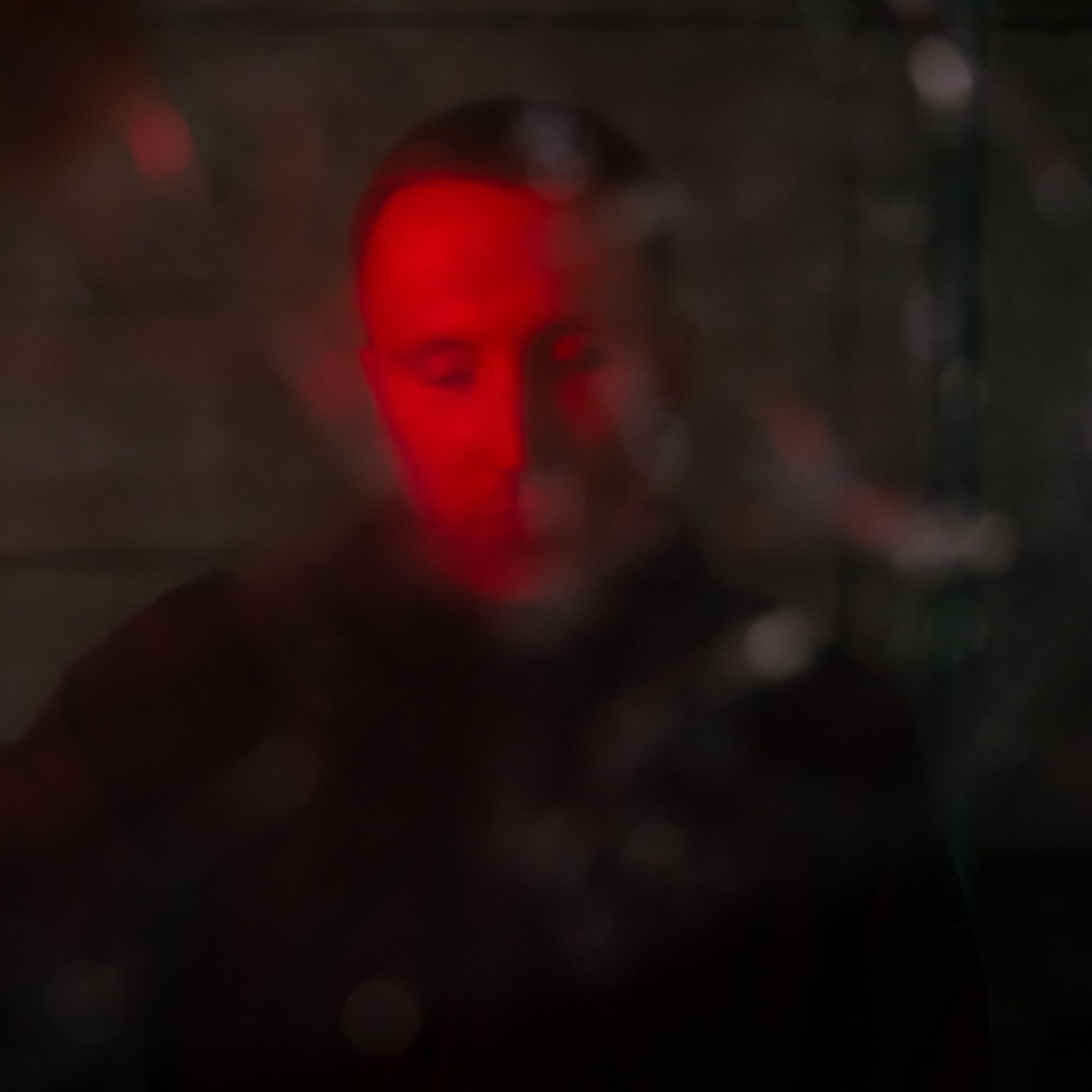 Das Foto zeigt den Rapper DrikOne. Er sieht nach unten und trägt eine schwarze Jacke. Das Foto ist in rot gehalten und hat ein verwaschenes Muster, das mit einer Folie vor dem Objektiv erzeugt wurde. Das Foto wurde von Fotograf Toni Borgert aufgenommen.