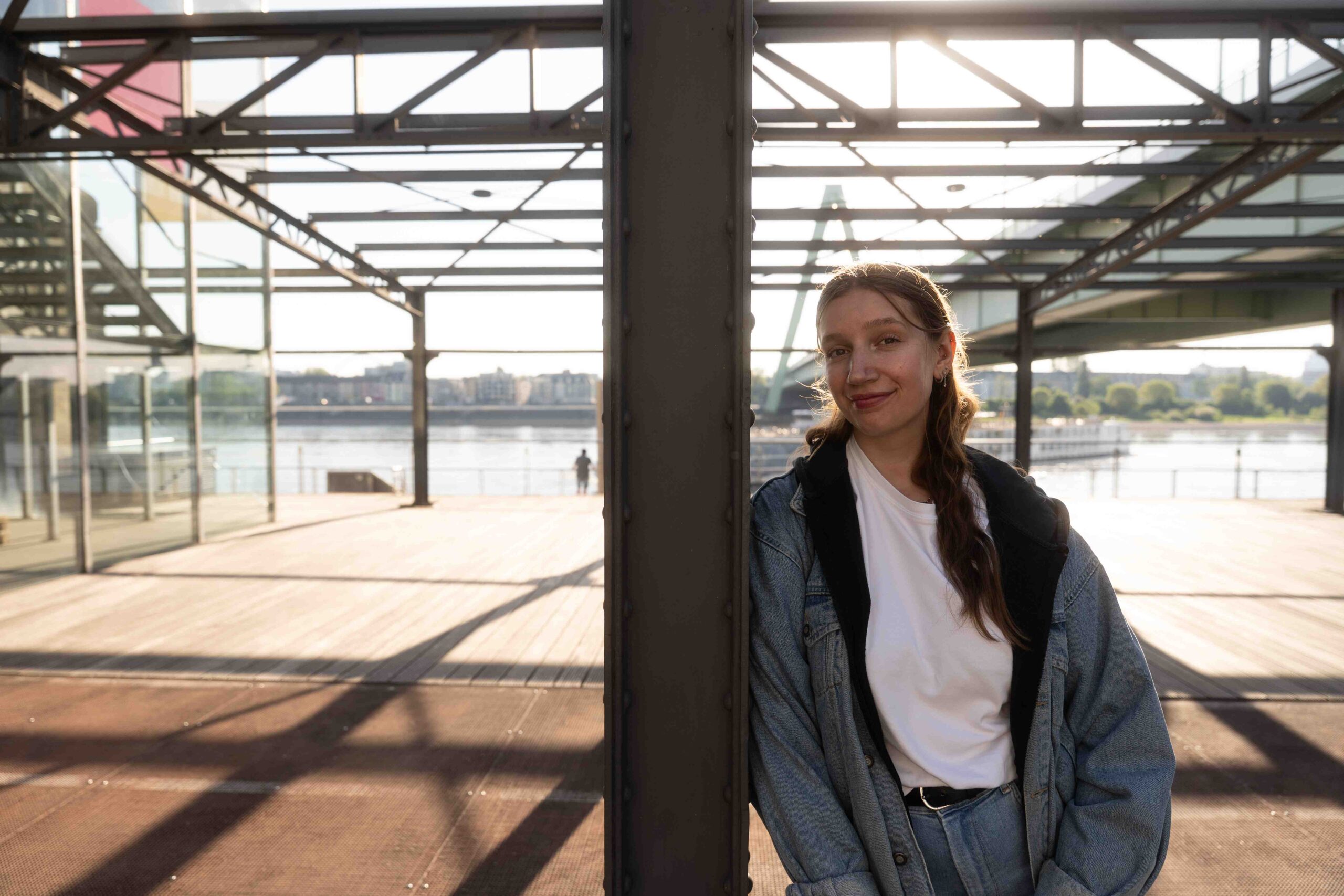 Das Bild zeigt eine junge Frau die unter einer Balkenkonstrultion steht. Die Konstruktion erinnert an ein Dach, ist aber lichtdurchlässig. Die Person steht seitlich an einem Stahlpfeiler und hat die Schulter angelehnt. Hinter ihr geht die Sonne auf und sieht steht im Gegenlicht. Im Hintergrund sieht man Teile des Rheinauhafens und der Severeinsbrücke in Köln. Die Frau trägt eine blaue Jeansjacke und eine passende Hose mit einem weißen T-Shirt. Sie hat schulterlanges blondes Haar und lächelt in die Kamera. Das Bild wurde von Fotograf Toni Borgert aufgenommen.