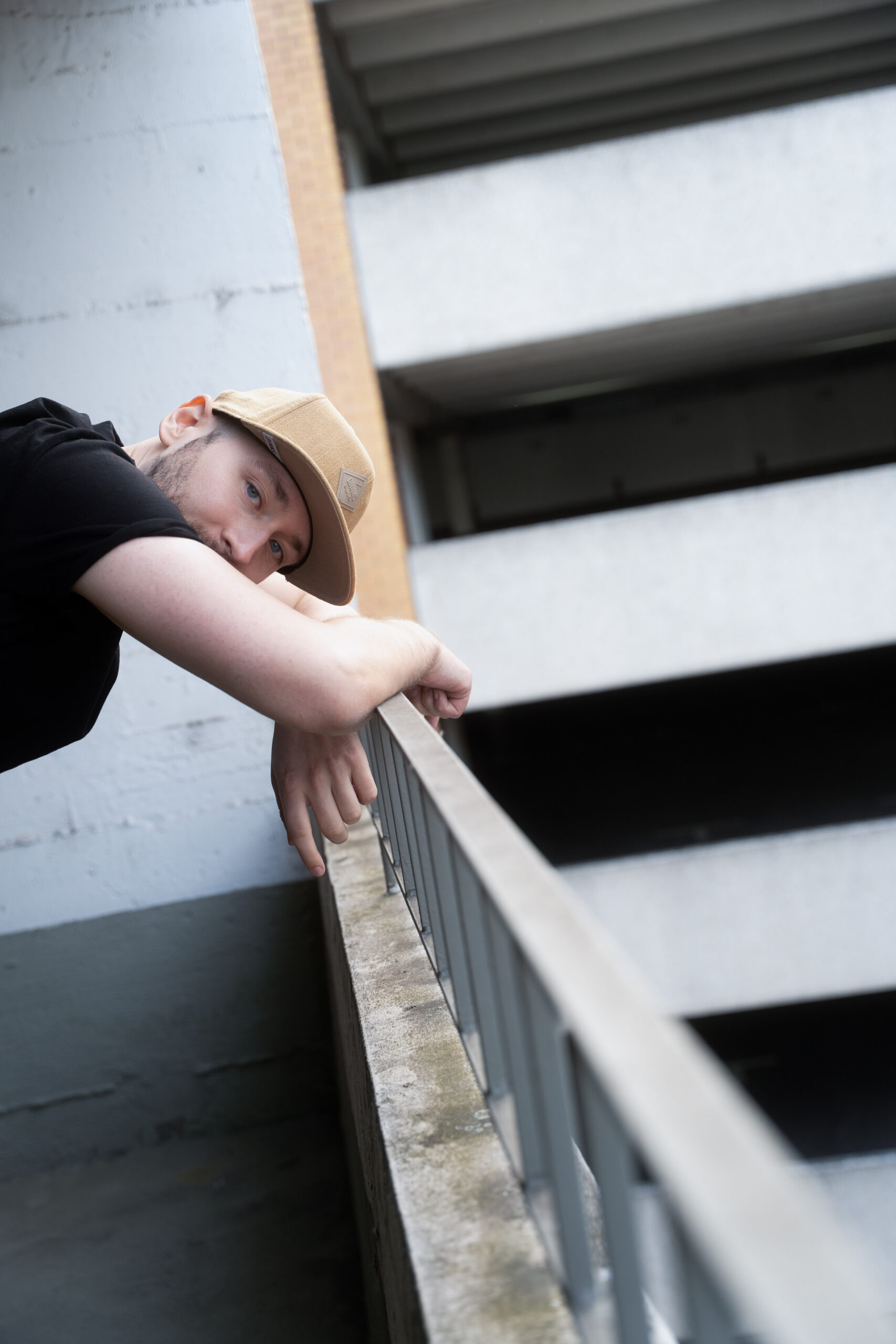 Das Bild zeigt eine männliche Person die mit den Armen nach vorne begeugt auf einem Geländer lehnt. Die Person trägt eine Baseballballkappe und ein schwarzes T-Shirt. Sie sieht seitlich in die Kamera, der Mund ist vom Arm bedeckt. Das Foto ist s o angeschnitten, dass nur der Kopf, die Arme und der Oberkörper zu sehen sind. Das Foto wurde von Fotograf Toni Borgert aufgenommen.