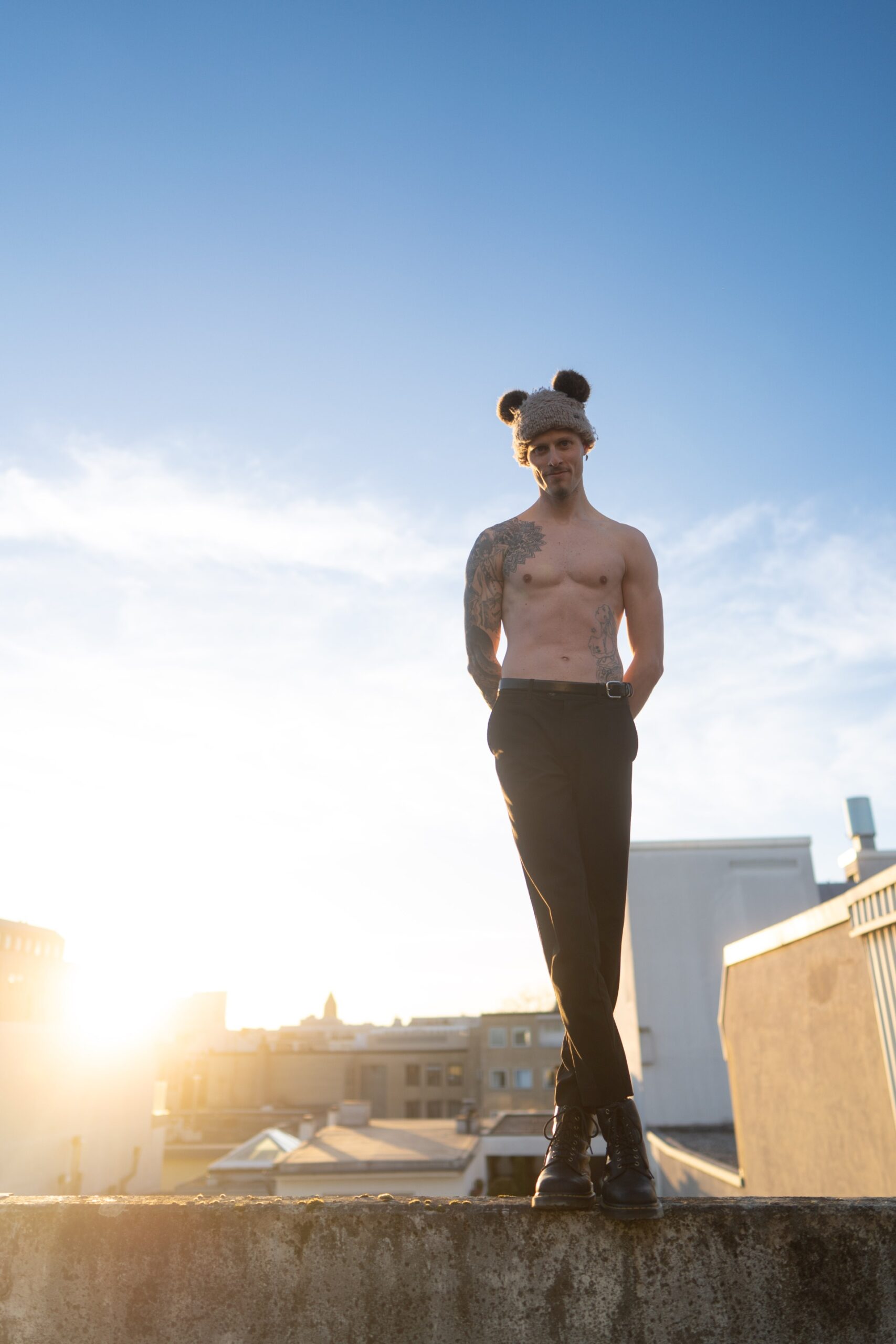 Auf dem Foto ist ein Mann zu sehen, der auf einer Mauer steht. Die Aufnahme zeigt seinen ganzen Körper. Er trägt eine Plüschmütze mit Ohren und kein Oberteil. Er hat eine schwarze lange Hose und Lederschuhe an. Er steht mit überkreuzten Beinen auf der Mauer und hat die Arme hinter dem Oberkörper verschränkt. An der linken Schulter hat er ein großes Tattoo bis auf den Oberarm. Das Foto wurde im Sonnenuntergang aufgenommen und zeigt blauen Himmel und eine diffuses Gegenlicht der Sonne. Das Foto wurde von Fotograf von Toni Borgert aufgenommen.