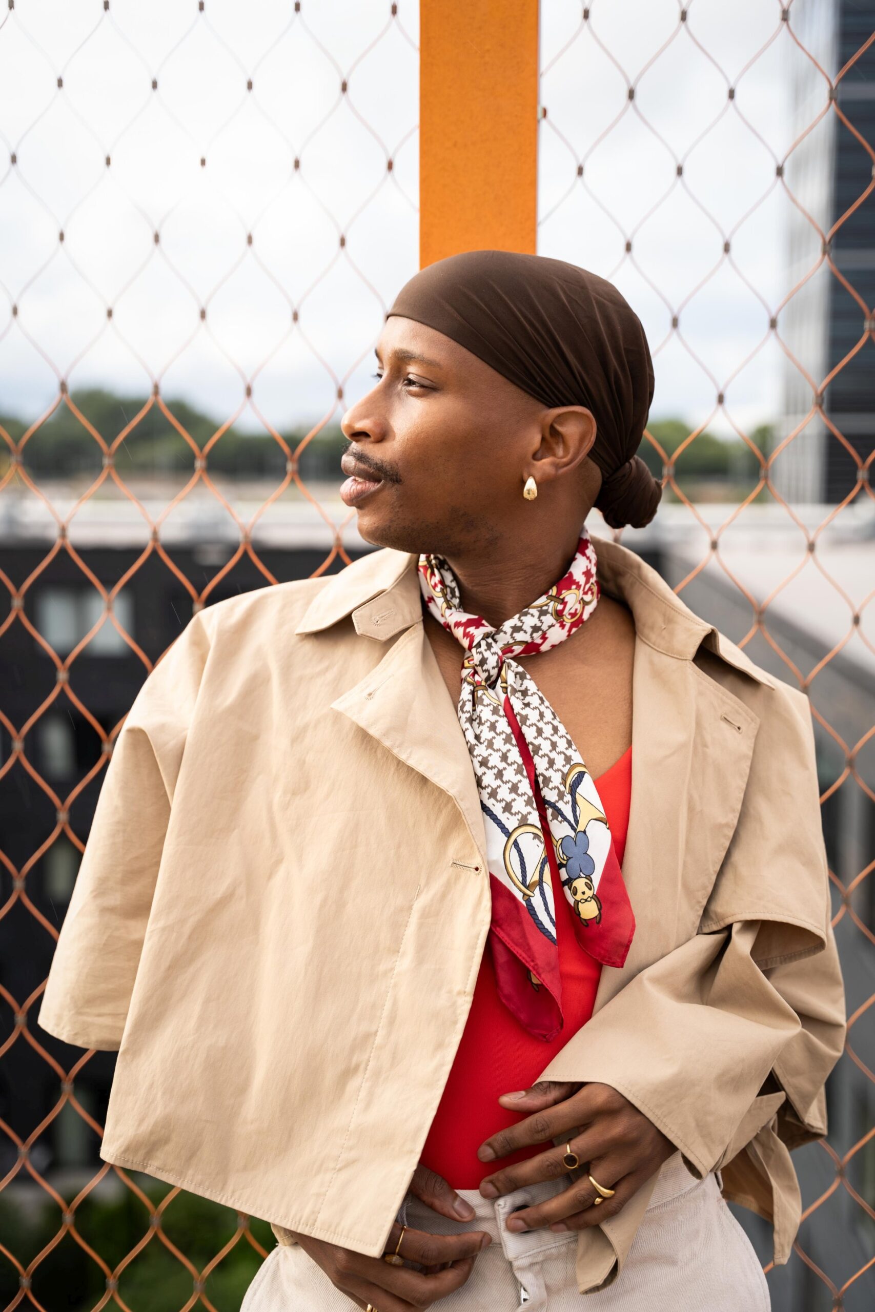 Auf dem Foto ist eine Person of Colour sehen. Sie sieht nach links, trägt einen Schnauzbart und ein braunes Kopftuch, das hinten zu einem Dutt gebunden ist. Die männlich gelesene Person trägt außerdem ein beiges Halstuch und ein beiges Cape- Darunter ist ein rotes Oberteil zu sehen. Die Person ist ein Gitter gelehnt, das im Hintergrund leicht zu erkennen ist. Die Fotografie sieht aus, wie Editorial-Mode-Fotoshooting. Das Foto wurde von Fotograf Toni Borgert aufgenommen.
