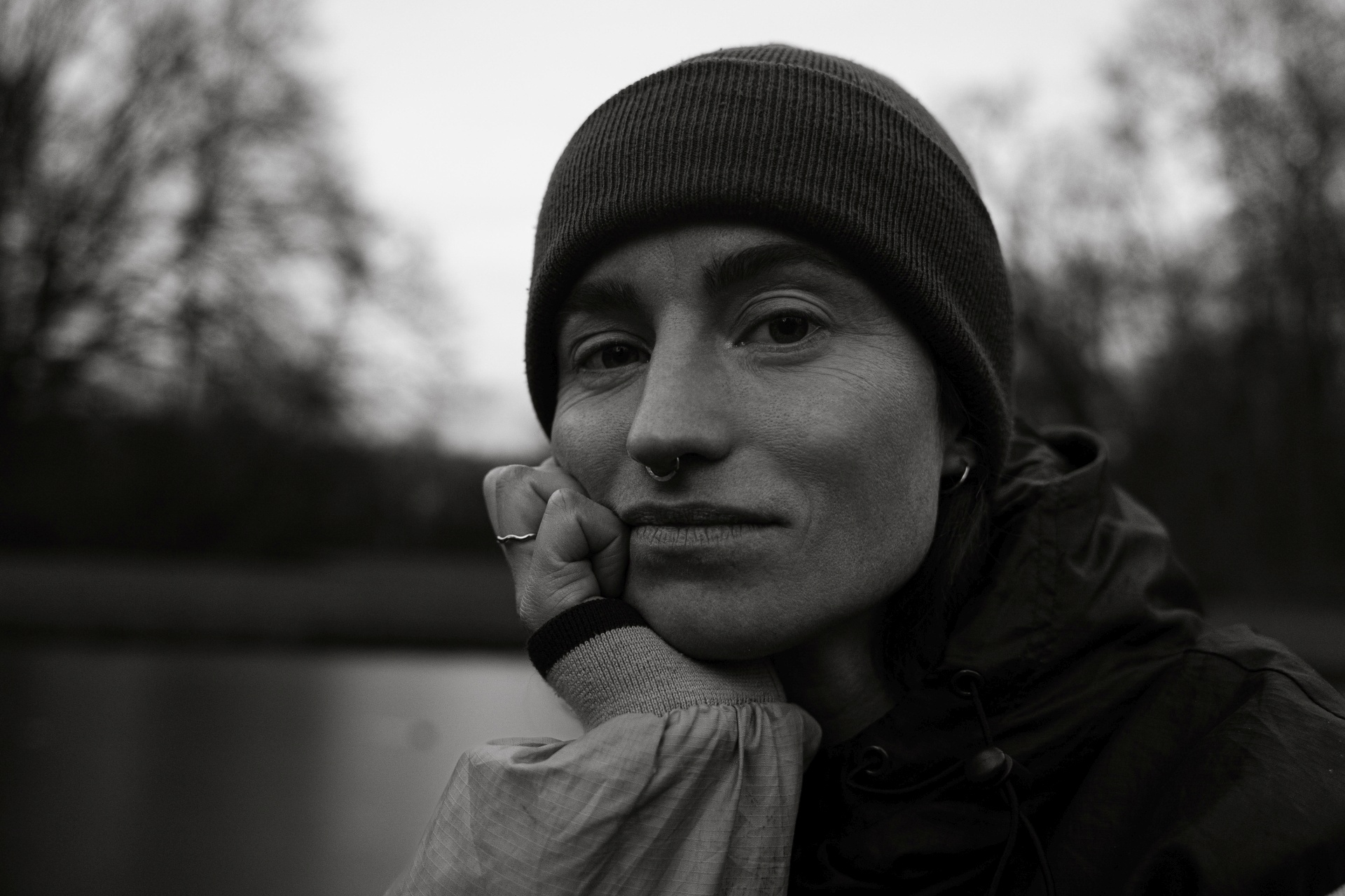 JanaR01-26_Copyright_TBorgert72 Das Foto zeigt eine weiblich gelesene Person. Zu sehen ist vor allem der Kopf und ein kleiner Teil des Oberkörpers. Die Person trägt eine Wollmütze und eine Trainingsjacke. Sie hat den Kopf auf der Hand abgestützt und sieht mit ernstem Gesichtsausdruck in die Kamera. Das Fotos schwarz-weiß aufgenommen. Das Foto hat Fotograf Toni Borgert fotografiert.