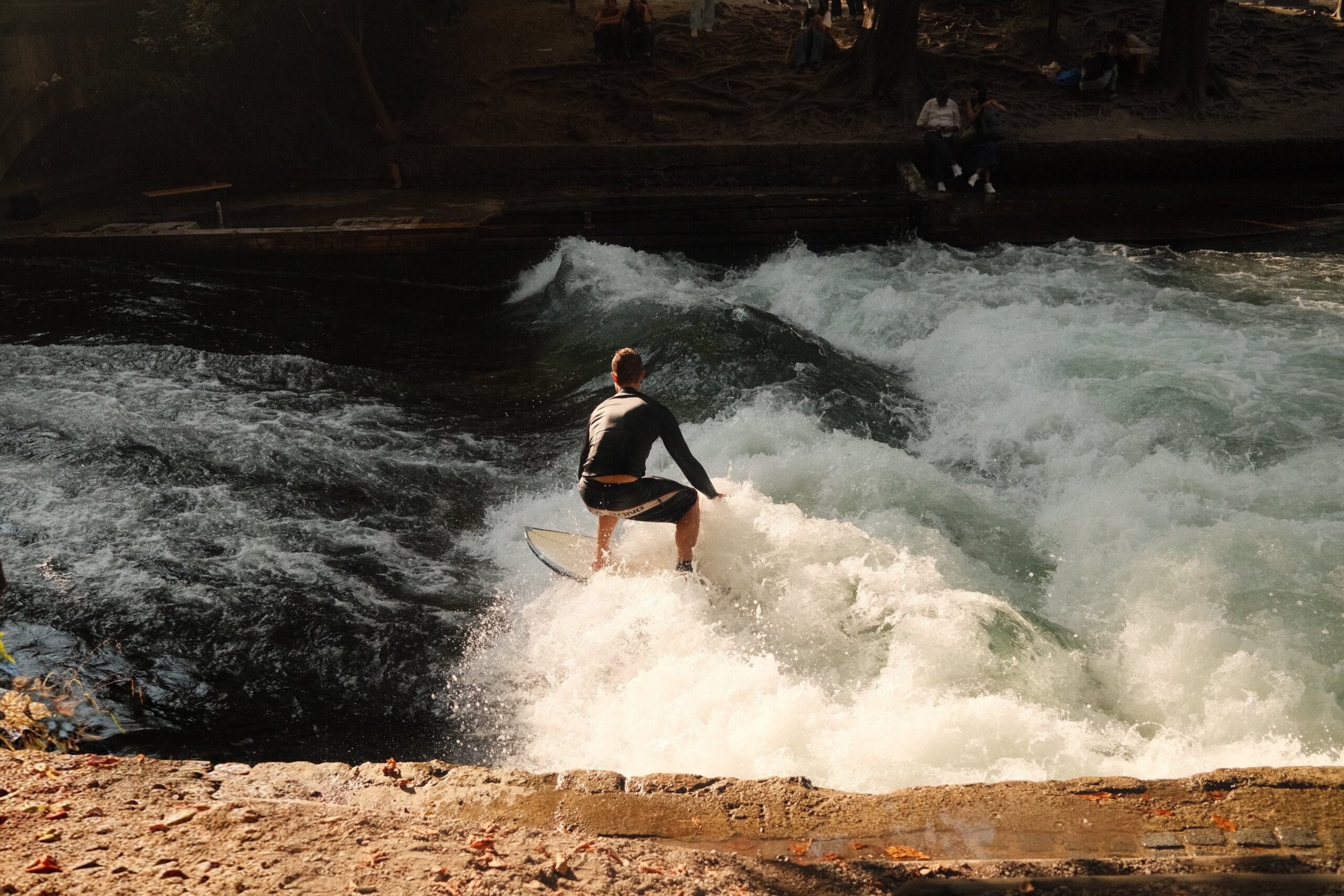 Muenchen_Surfe_Eisbach_©TBorgert Das Foto zeigt den Wasserfall am Eisbach in München. Es ist ein sonniger Tag, seitlich fällt Sonne auf das rauschende Wasser. Auf dem Wasser ist ein Surfer in dunkler Surferkleidung auf seinem Surfbrett zu sehen. Im Vordergrund erkennt man die Mauer, die den Bach begrenzt. Das Foto wurde von Fotograf Toni Borgert aufgenommen.