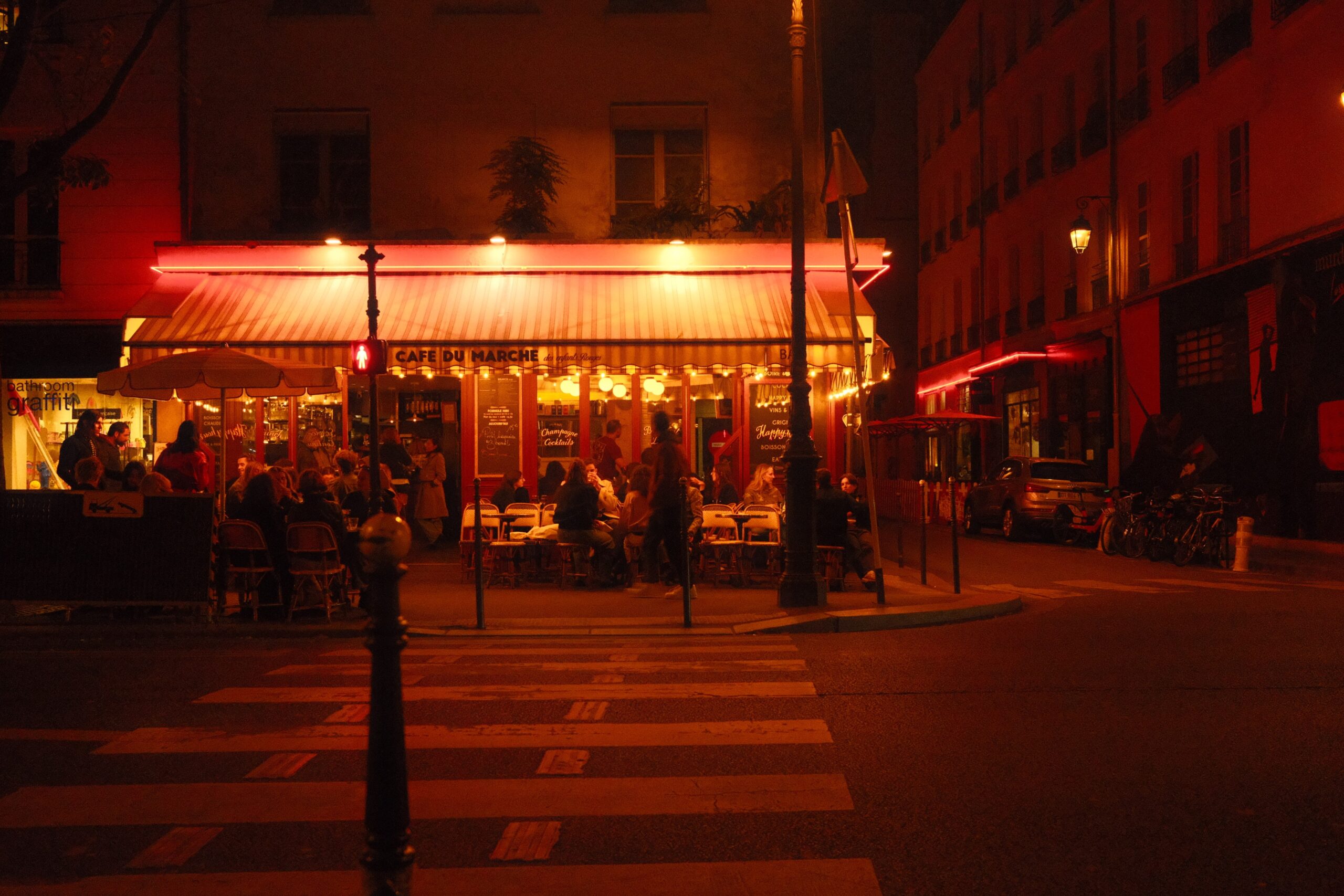 Paris_Abends_Strassencafe©TBorgert Das Foto zeigt eine Straße in Paris bei Nacht. Im Hintergrund ist ein Café mit Markise und Beleuchtung zu sehen. Davor sitzen Menschen und es ist ein Zebrastreifen mit Ampel zu sehen. Das Foto wurde von Fotograf Toni Borgert aufgenommen.