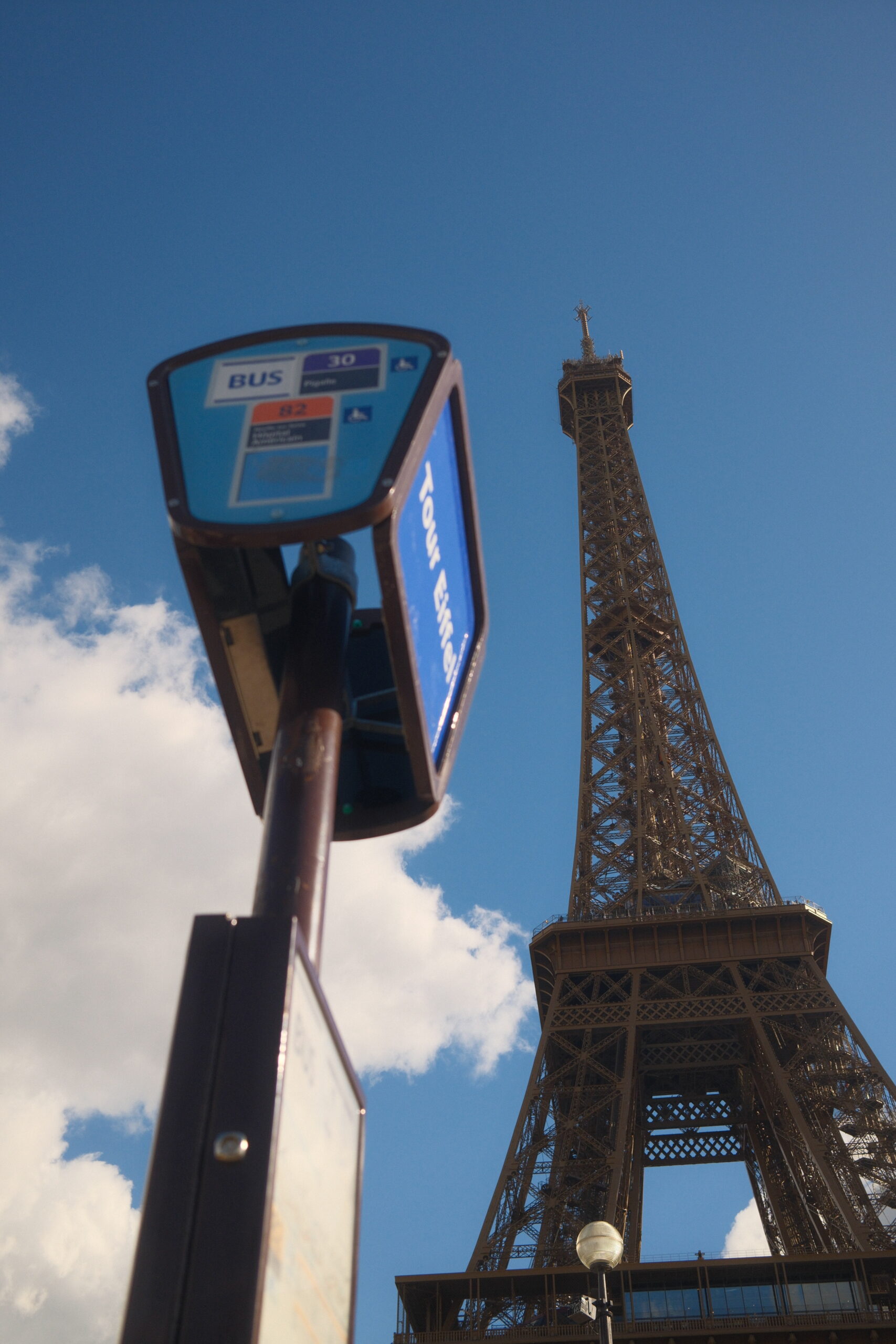 Paris_Eiffelturm_mit_Schild Das Foto zeigt einen Teil des Eiffelturms in Paris. Das Foto wurde sehr nah und steil nach oben fotografiert. Es ist ein großer Teil des Eiffelturms von etwa der Mitte bis zur Spitze zu sehen. Links daneben ein Haltestellenschild mit dem Namen der Haltestelle. Im Hintergrund sind Wolken und blauer Himmel zu sehen. Das Foto wurde von Fotograf Toni Borgert aufgenommen.