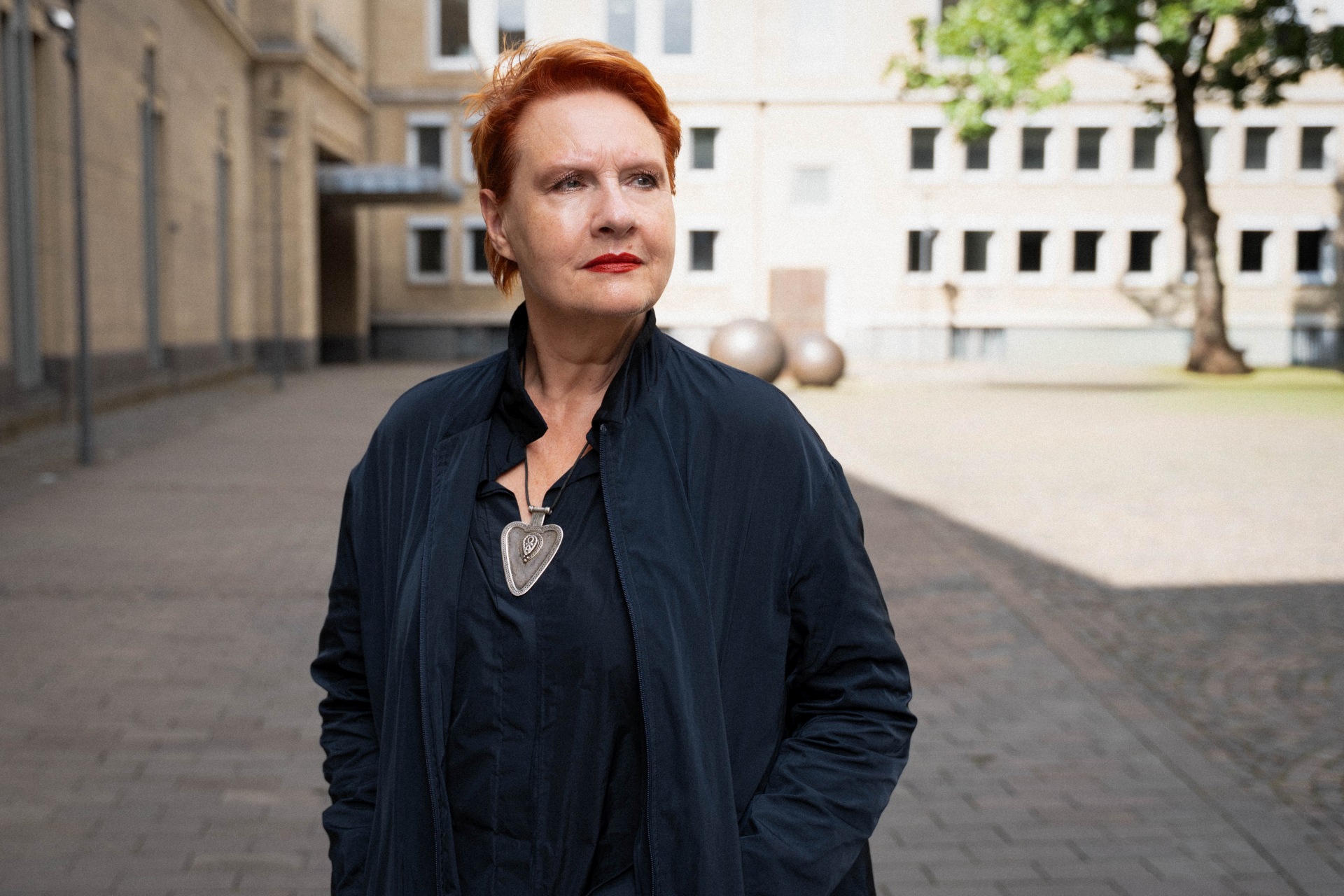 Schauspielerin_AnkeZillich_Copyright_TBorgert06 Das Foto zeigt die Schauspielerin Anke Zillich. Sie hat rote Haare und trägt eine blaue Jacke und ein schwarzes Oberteil. Über dem Oberteil ist eine Kette zu sehen mit einem großen Anhänger. Anke Zillich hat die Hände in den Taschen der Jacke und sieht leicht nach rechts. im Hitergrund ist eine Art Hinterhof und ein Gebäude zu sehen. Die Sonne scheint, die Schauspielerin steht im Schatten.