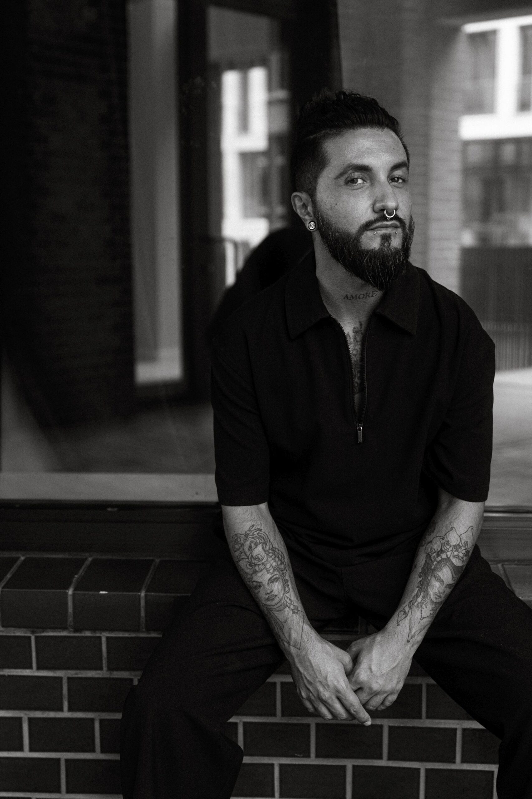 StefandP2025_Copyright_TBorgert92 Das Foto zeigt einen Mann, der auf einem Fenstersims sitzt. Er trägt ein schwarzes Oberteil und schwarze Hosen. Sein Oberteil hat kurze Ärmel und seine Tattoos auf den Armen sind zu sehen. Er hat lange schwarze Haare die zu einem Dutt zusammengebunden sind. Er trägt einen Bart und sieht leicht nach rechts, aber mit den Augen in die Kamera. Er sitzt leicht nach vorne gebeugt und hat die Arme nach unten fallen lassen. Eine Hand greift dabei die andere Hand. Es ist ein Stück geflieste Wand zu erkennen. Das Foto ist schwarz-weiß und wurde von Toni Borgert aufgenommen.