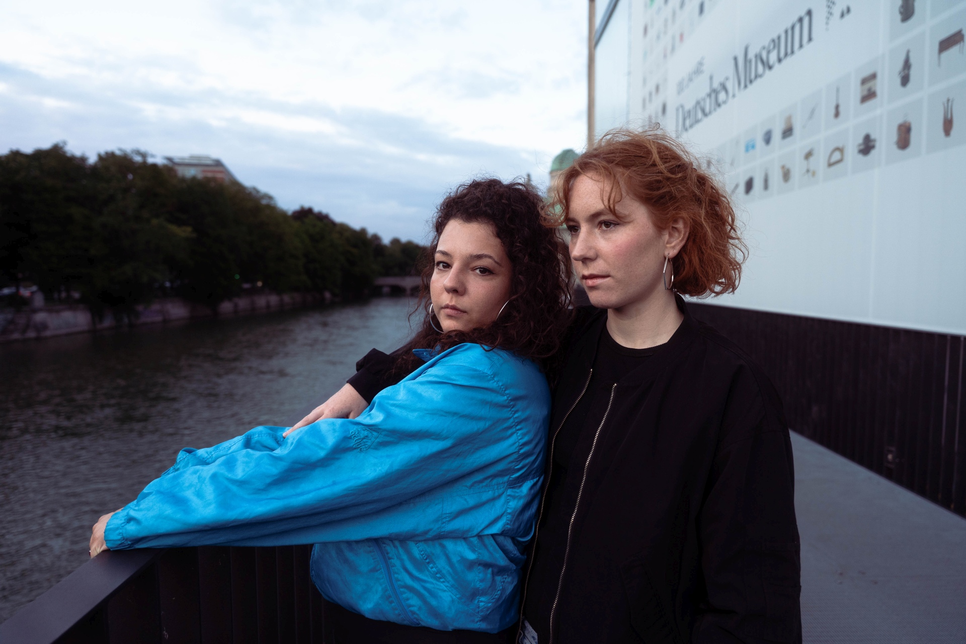 Auf dem Foto sind zwei Frauen zu sehen. Die Linke Frau hat schwarze lockige Haare und trägt eine hellblaue Jacke aus Ballonseide. Die Frau rechts hat rote lockige Haare, die bis zum Ohr reichen. Sie trägt eine schwarze Jacke. Die Frau in Blau hält sich mit beiden Händen an einem Geländer fest. Die andere steht hinter ihr und hat einen Arm um ihre rechte Schulter gelegt. Die Linke sieht in die Kamera, während die Rechte in die Ferne blickt. Beide stehen auf einer Brücke am Geländer. Das Foto wurde von Fotograf Toni Borgert aufgenommen.